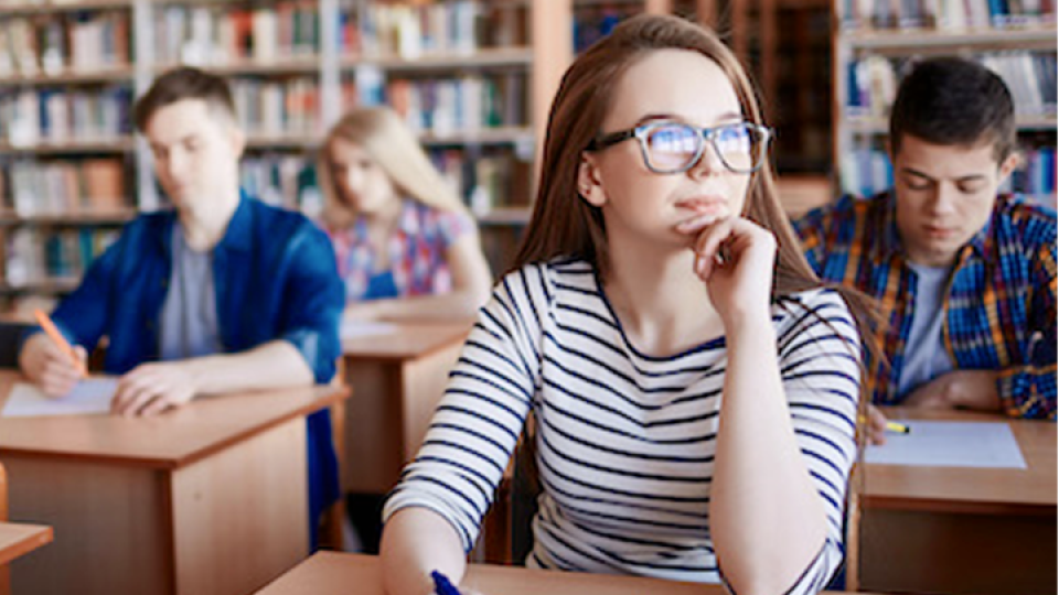 Ragazzi a scuola seduti nei banchi. In primo piano c'è una ragazza sorridente con in mano una penna.